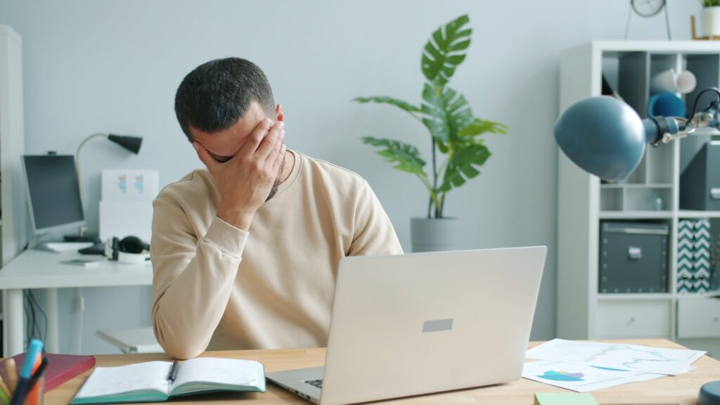 Man rubbing his eyes in front of a laptop computer screen, showing digital eye strain